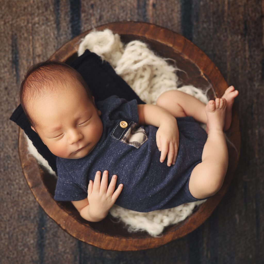 Sleeping newborn boy in a blue onesie curled up in a wooden bowl photographed from above