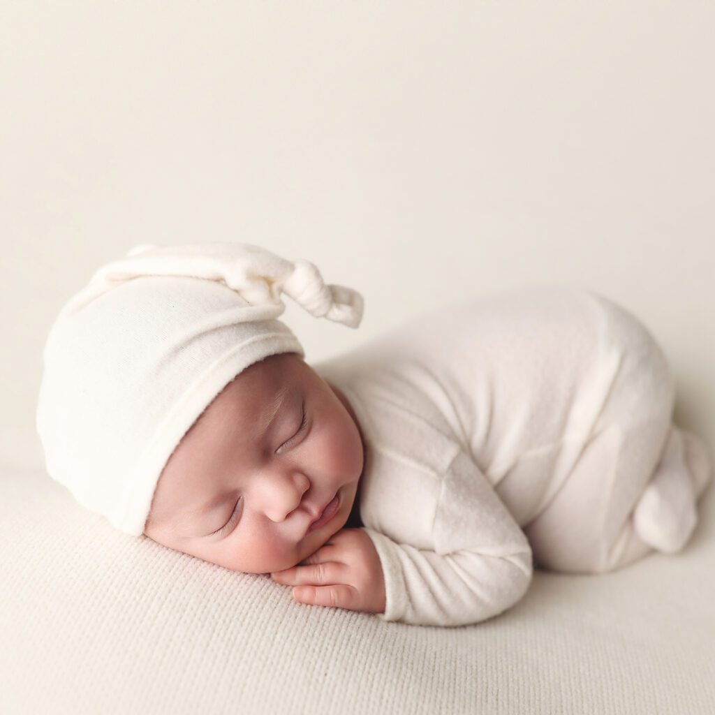 Sleeping newborn baby boy wearing a white romper and cap lying on his tummy
