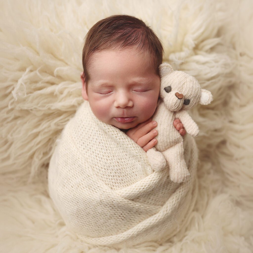 Newborn baby boy swaddled in white holding a small white teddy bear on a fur backdrop