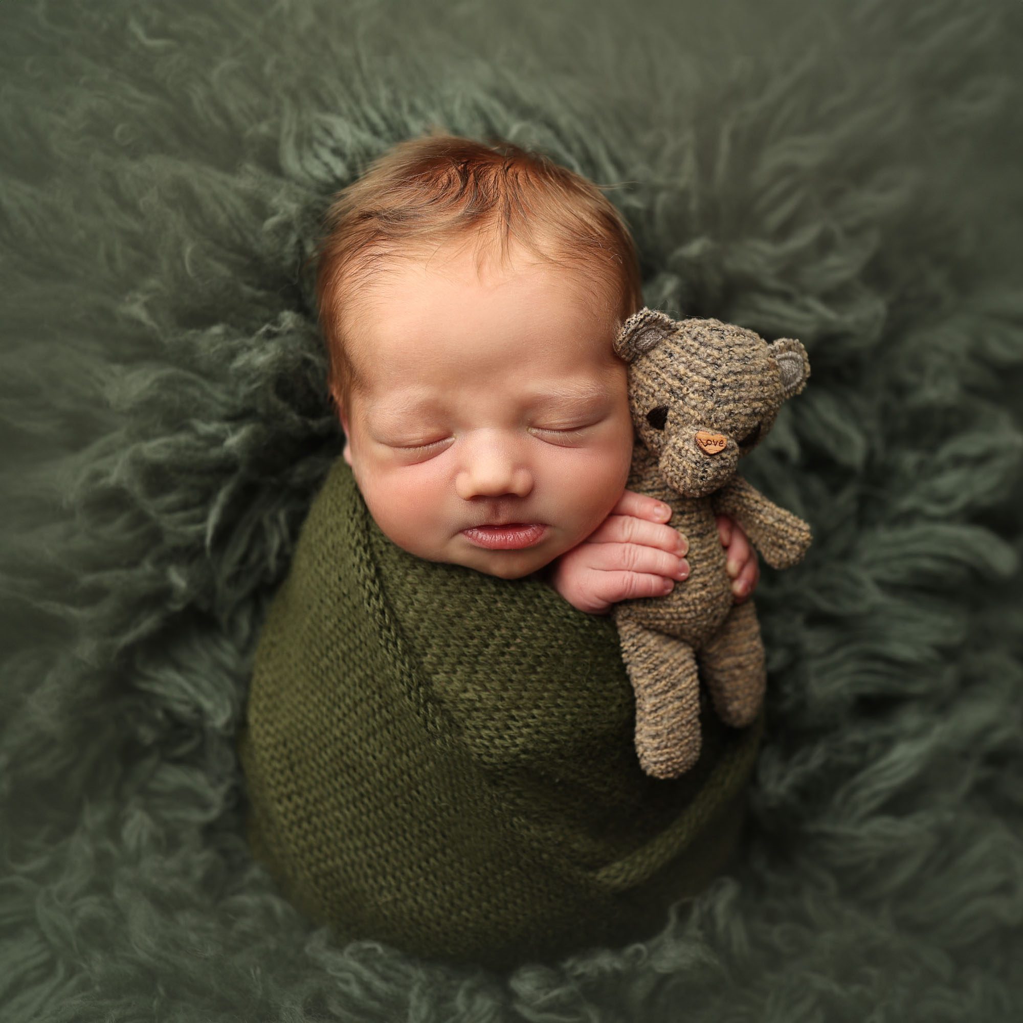 Liberty, Mo newborn baby boy in dark green knit wrap holding small brown teddy bear on fur rug