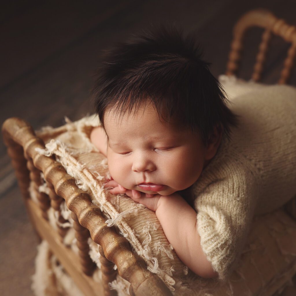 Newborn baby boy with thick hair sleeping on a wooden bed with his chin resting on his hands