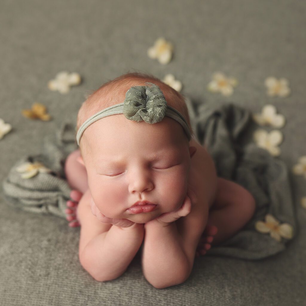 Newborn baby girl in froggy pose on sage green fabric with yellow flower petals