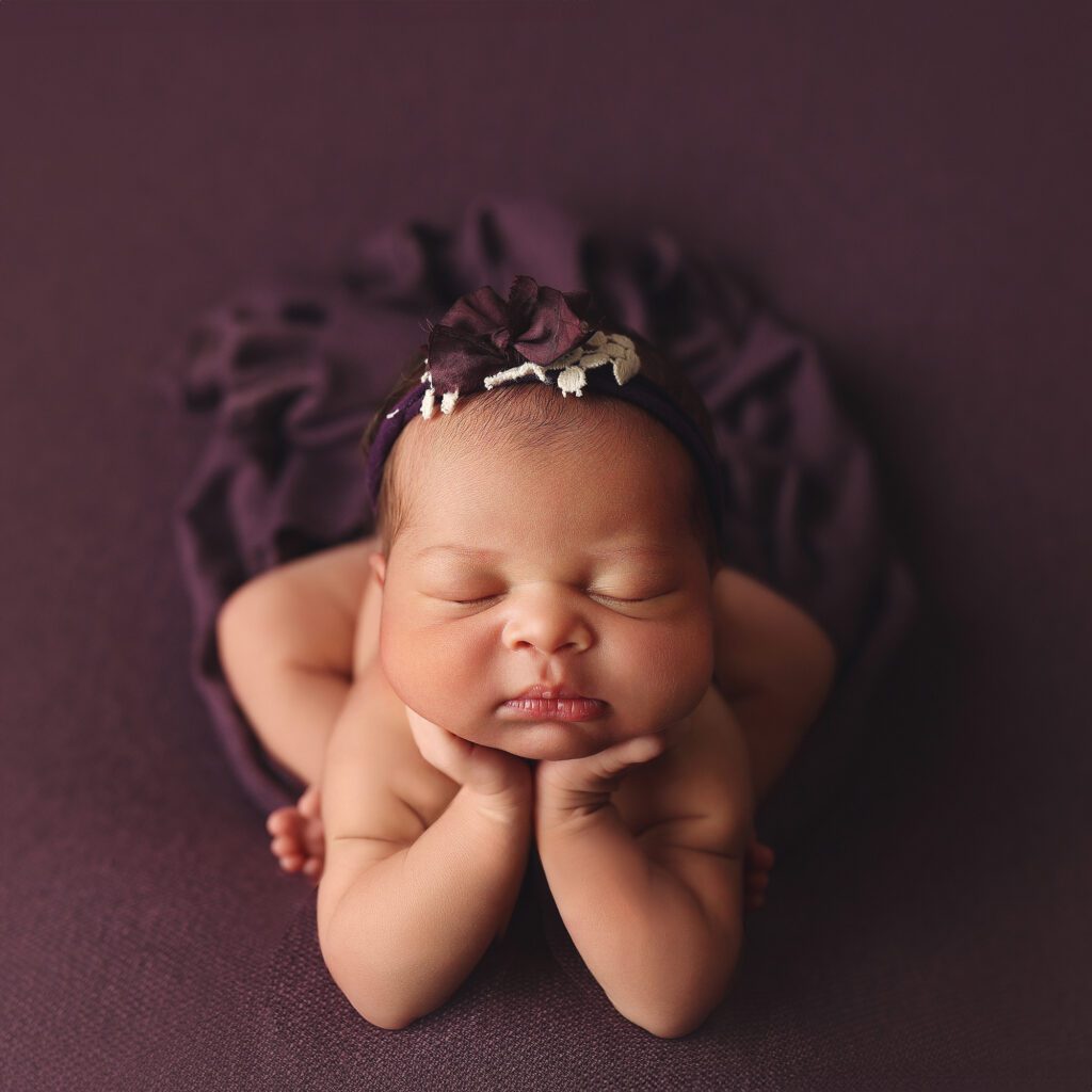 Newborn baby girl in froggy pose on dark plum fabric with a trailing wrap