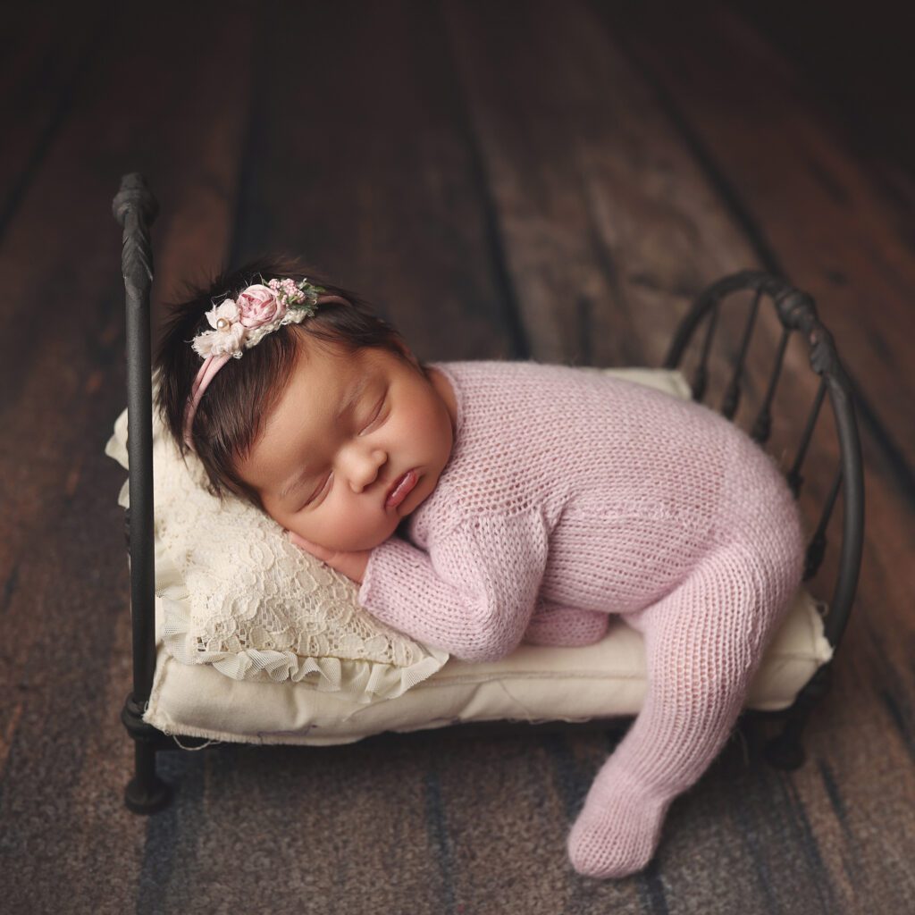 Newborn baby girl wearing a pink knit sleeper on a small wrought iron bed