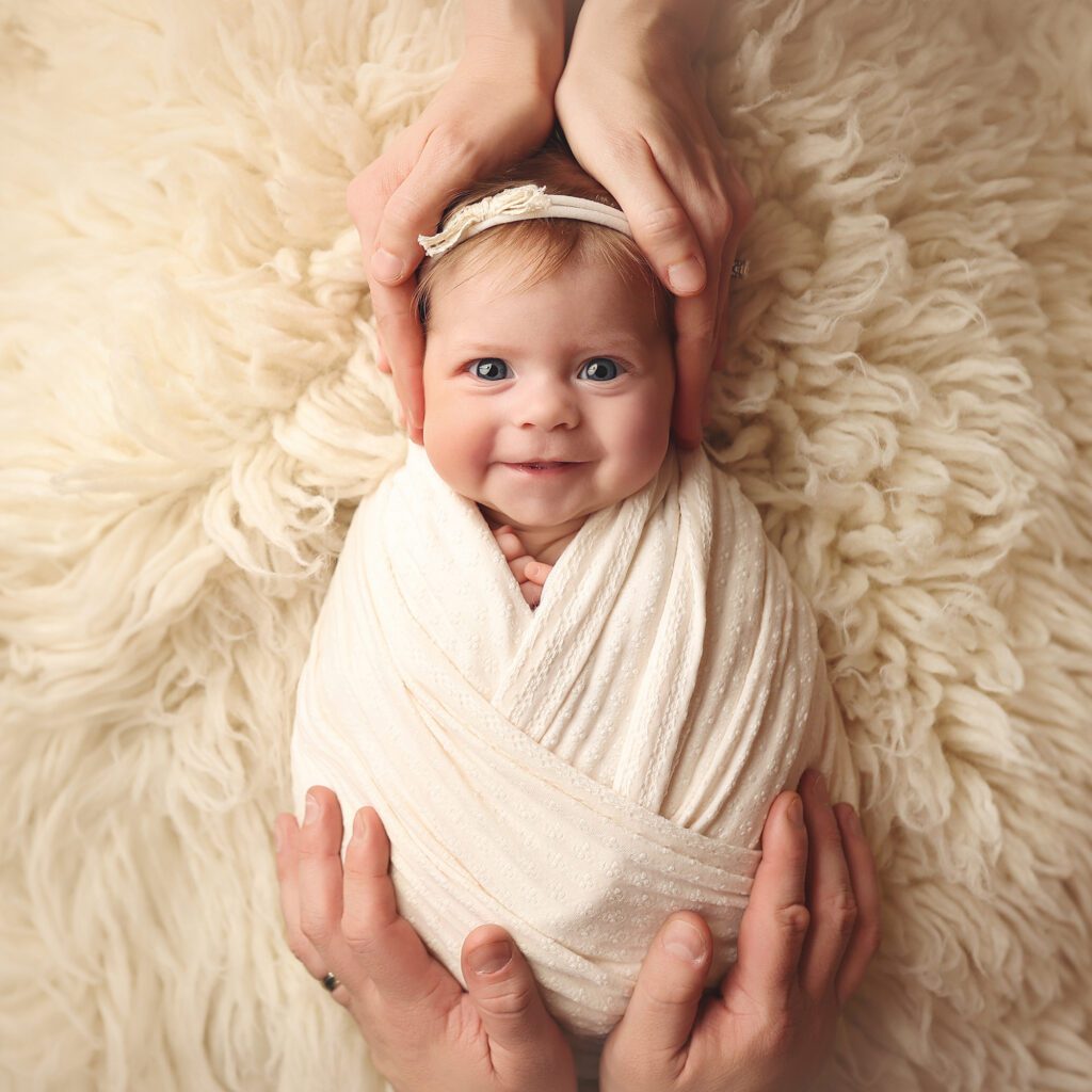 newborn baby smiling at the camera with parents' hands gently resting on her head and body