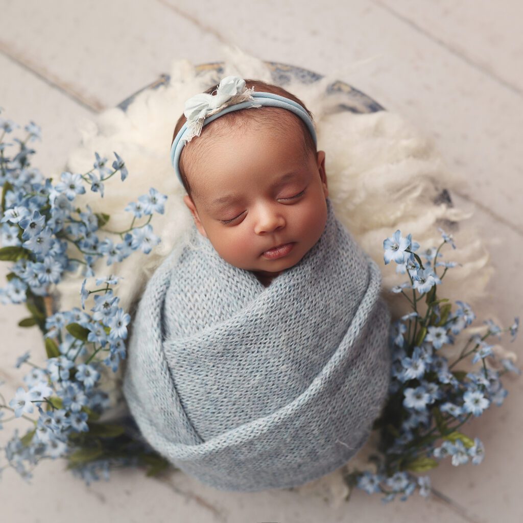 Newborn baby girl wrapped in blue knit fabric in a white bowl with blue flowers photographed from above