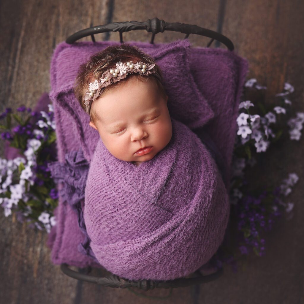 newborn baby girl wrapped in purple on a wrought iron bed surrounded by purple flowers