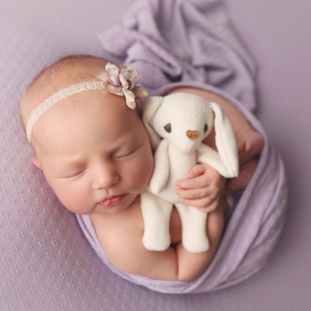 newborn baby girl wrapped in lavender holding a small white stuffed bunny