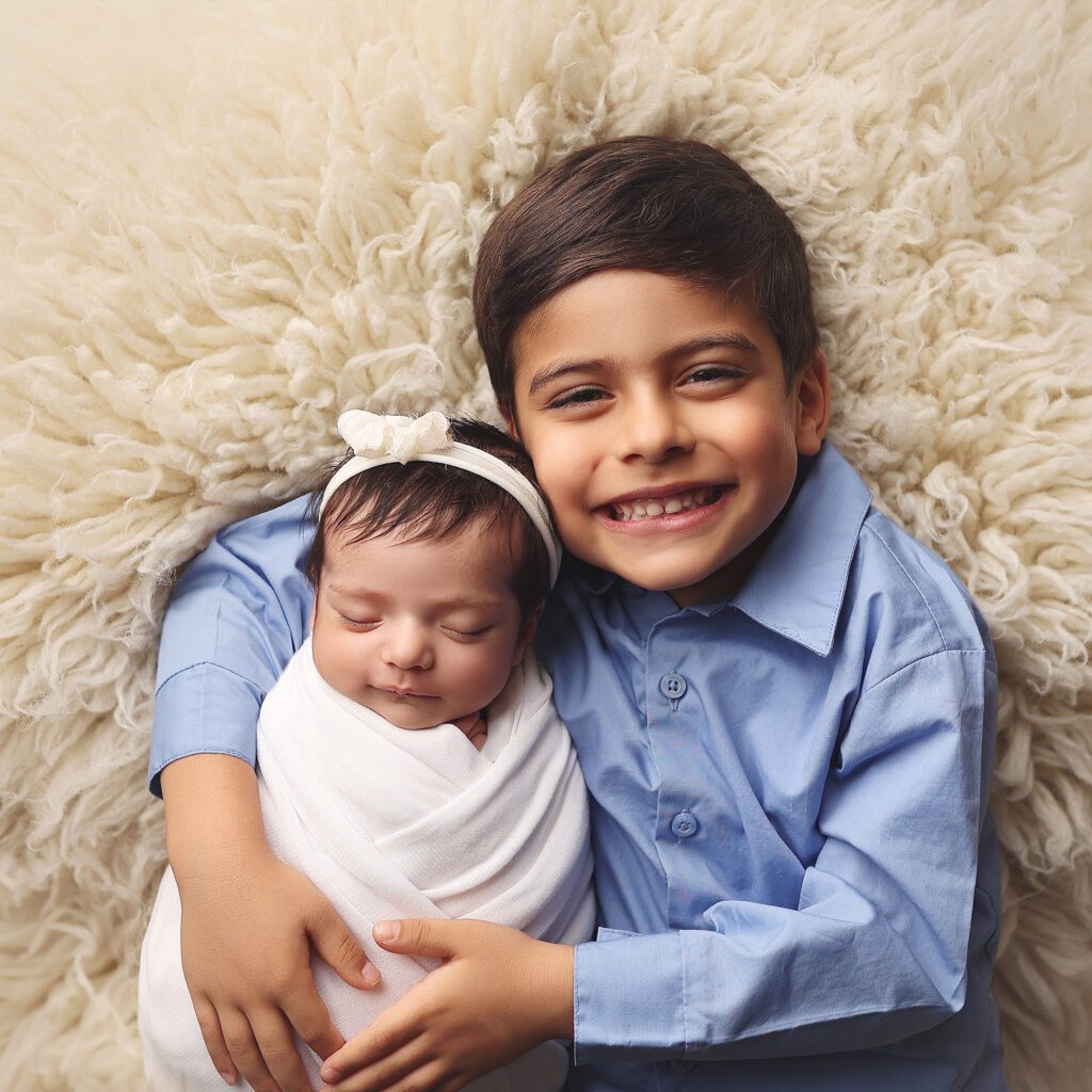 Older brother smiling at the camera while holding his newborn sister wrapped in white