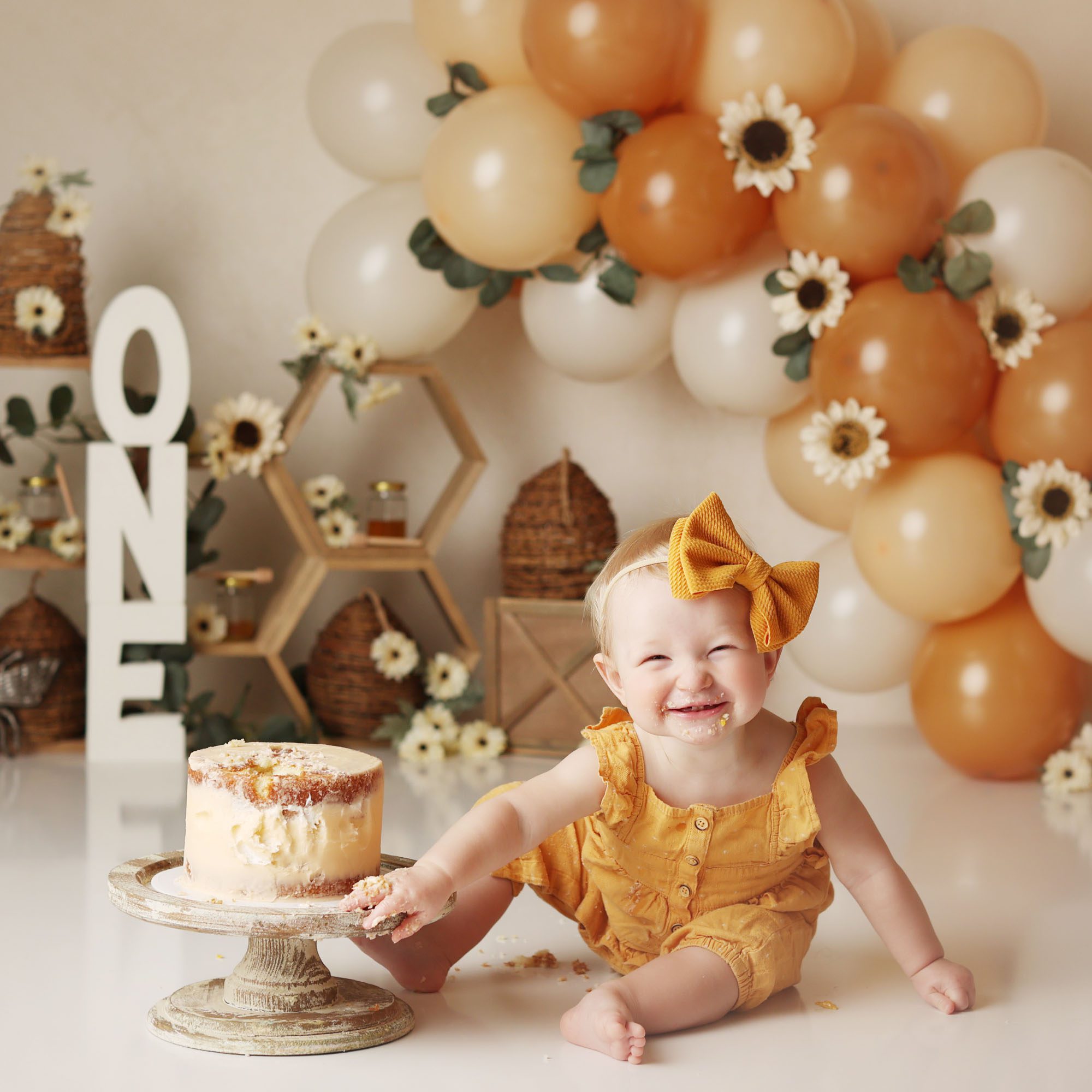 Blue Springs, MO Honeybee cake smash girl in gold romper with beehvie decor and jars of honey smiling at the camera