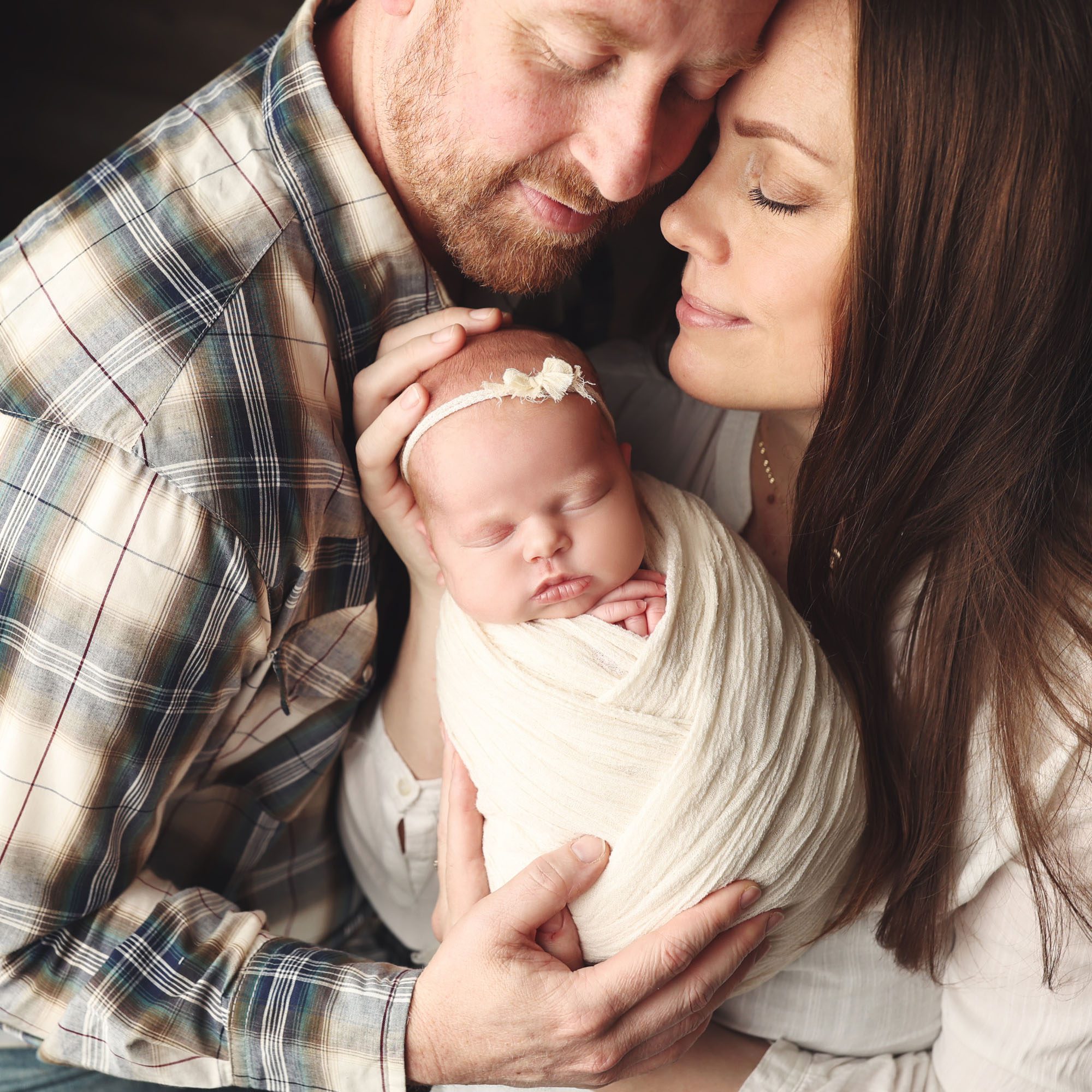 Lee's Summit, MO parents holding newborn baby girl wrapped in white with bow in a close up studio portrait