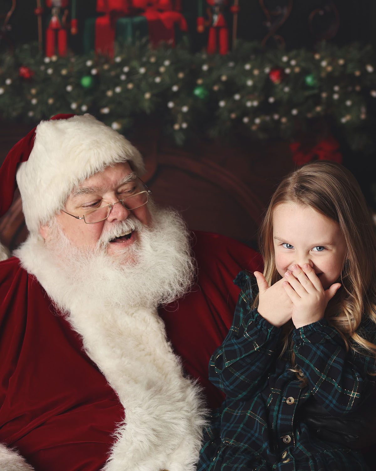 Santa laughing with a little girl in a plaid dress as she smiles at the camera during a Christmas session